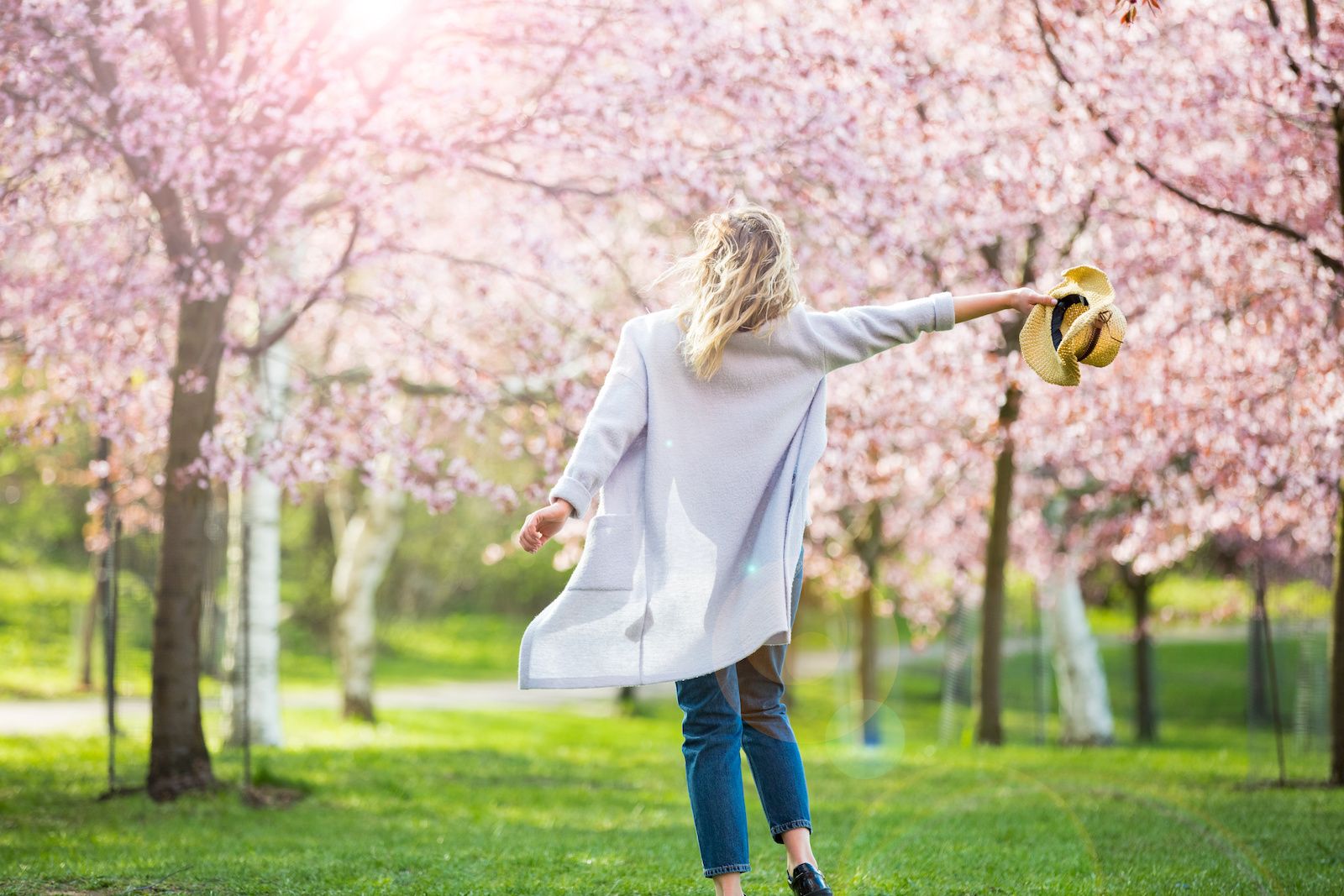 Woman walking on grass through beautiful blooming trees with pink blooms while holding her hat out to the side