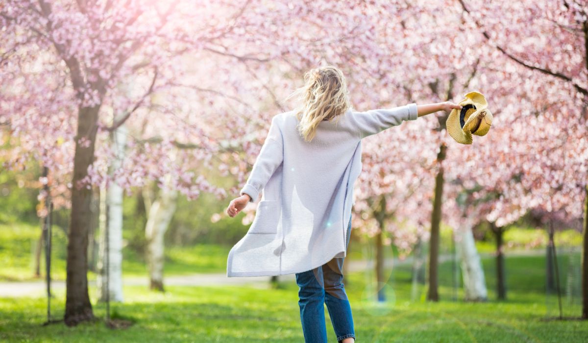 Woman walking on grass through beautiful blooming trees with pink blooms while holding her hat out to the side
