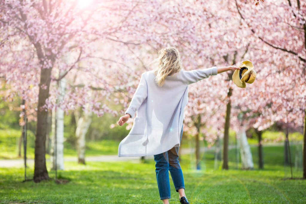Woman walking on grass through beautiful blooming trees with pink blooms while holding her hat out to the side
